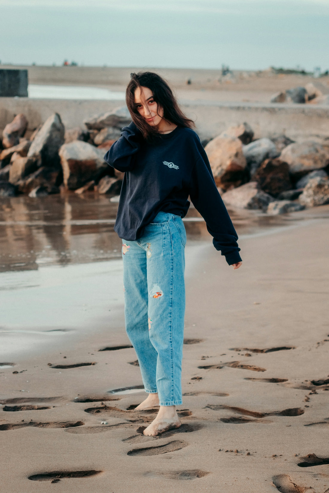 woman in black long sleeve shirt and blue denim jeans standing on beach shore during daytime